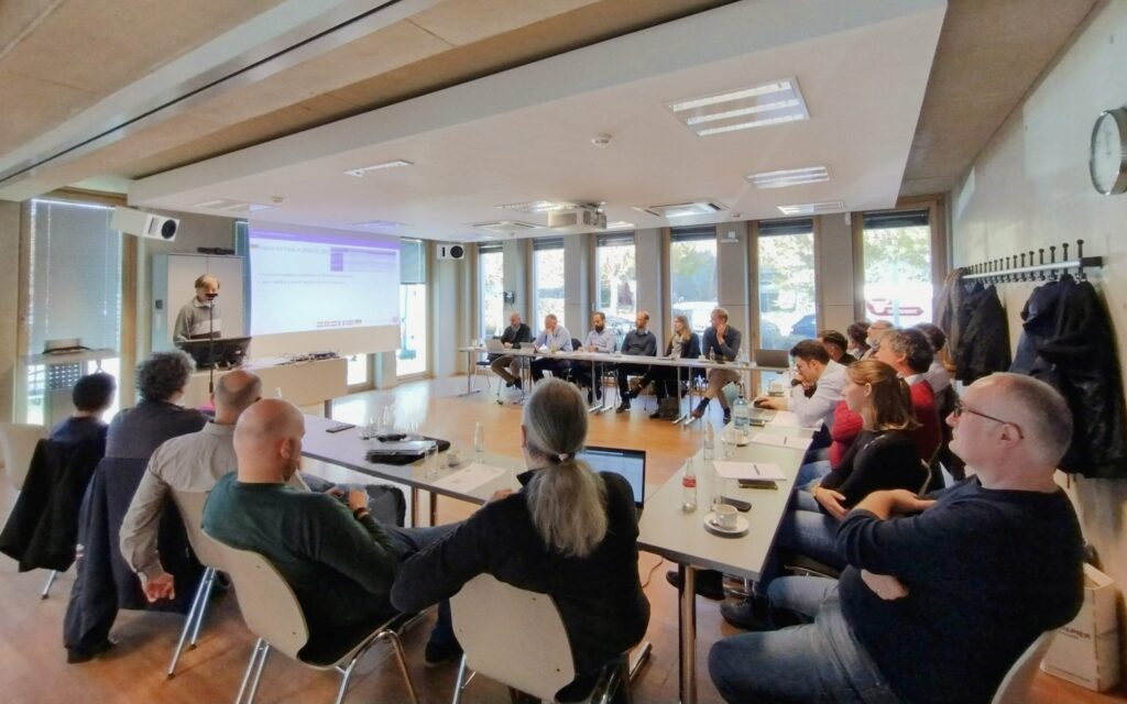 The photo shows the participants of the IEA HPT Annex 63 meeting in Aachen sitting in the conference room of the Institute for Hearing Technology and Acoustics, RWTH Aachen University.