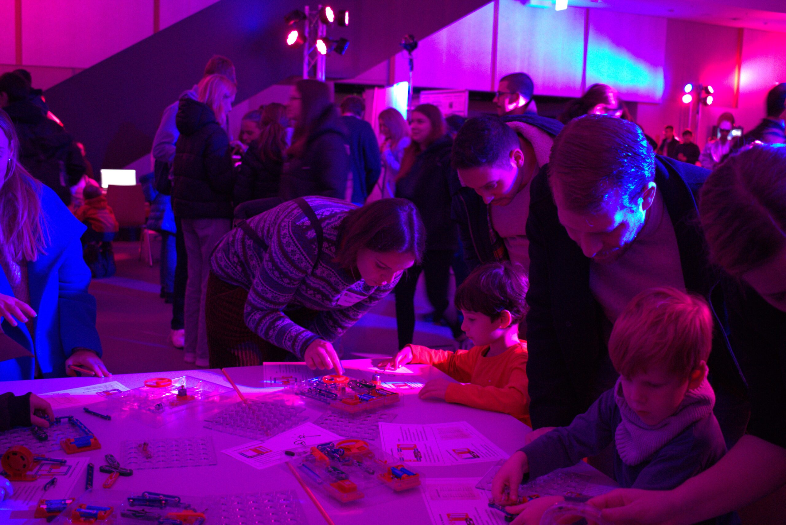 Children sit at a table equipped with electronic construction kits and are guided by adults. In the background, other people are on the move in the event room.