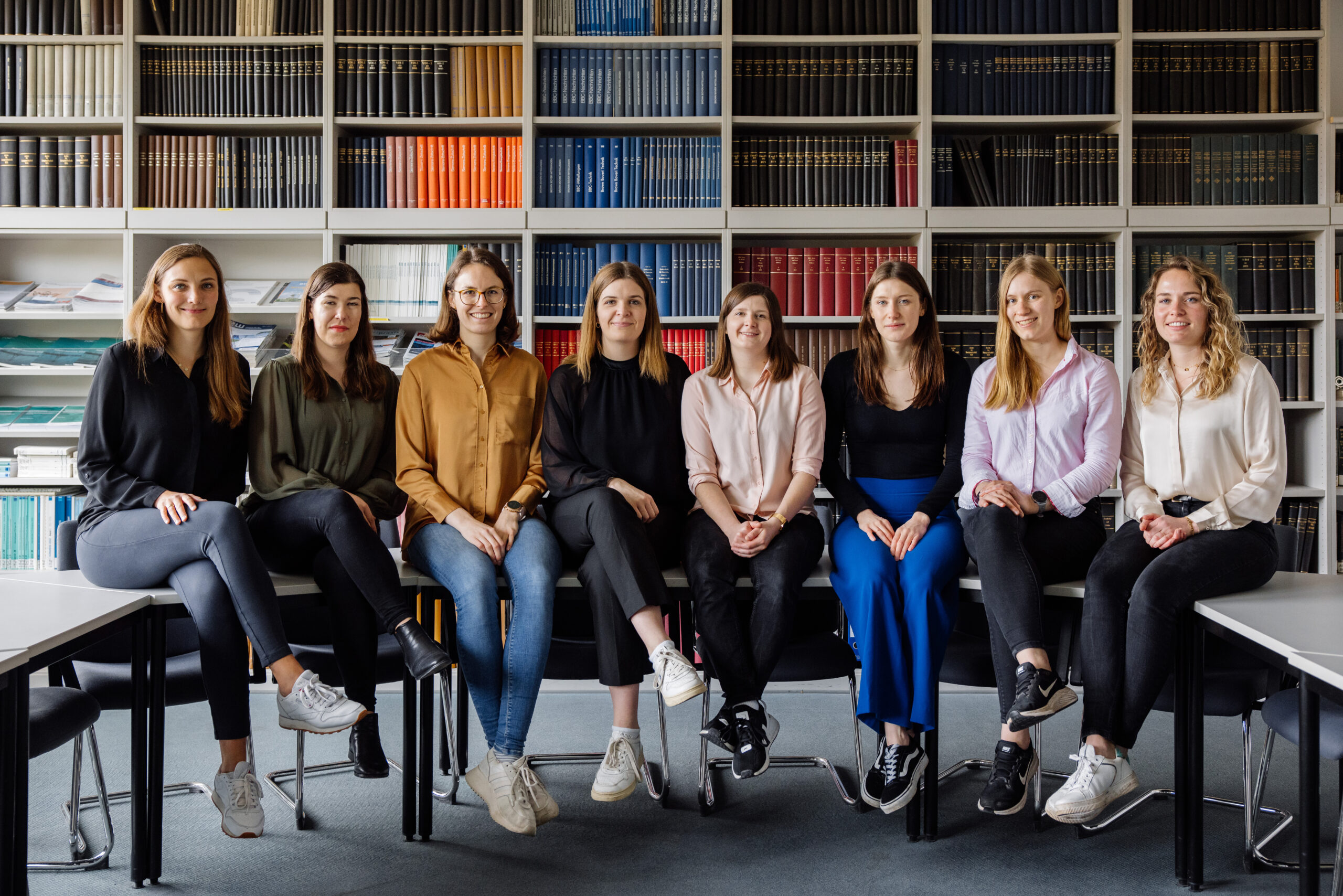 Sitting in a row, eight young women present themselves in front of a large bookshelf that takes up the entire wall behind them.