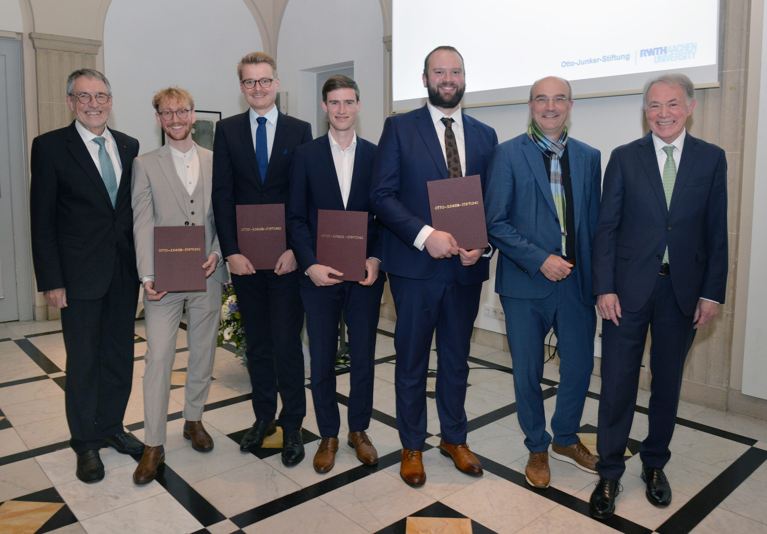 The formation is completed on the left by an elderly gentleman and on the right by two other elderly gentlemen; in the centre are four young men, each holding a certificate. The depicted individuals exhibit a smile directed towards the camera.