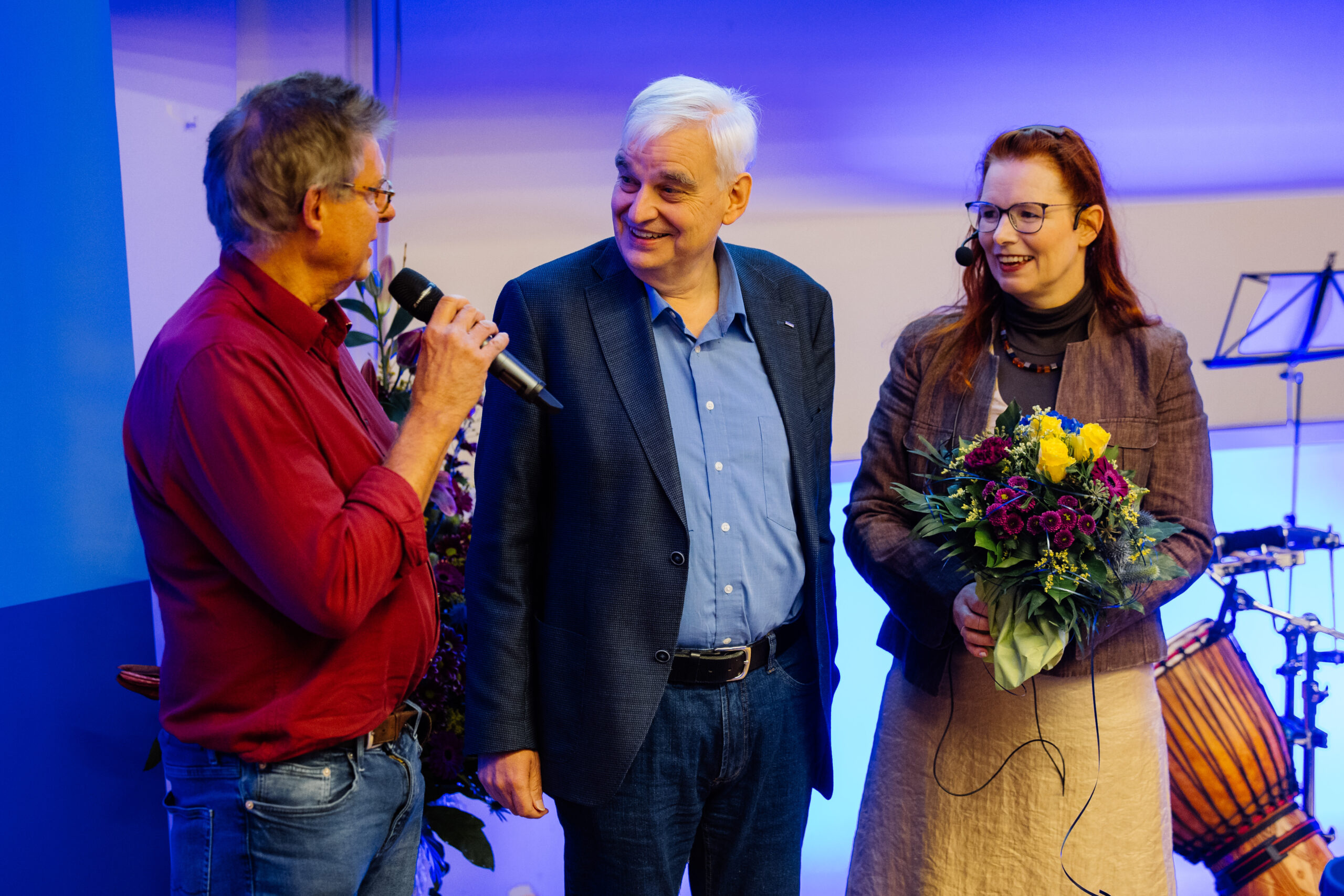 Auf der Bühne stehen eine Frau und zwei Männer. Die Frau, die einen Blumenstrauß trägt, und der Mann, der neben ihr steht, richten ihre Blicke auf die dritte Person, die ihnen zugewandt ein Mikrofon hält. 