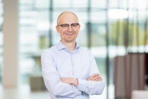 Portraitaufnahme eines lächelnden Mannes im hellblauen Hemd mit verschränkten Armen vor der Brust.