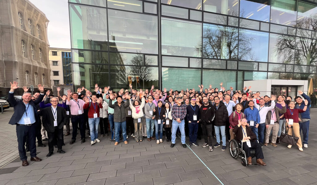 Eine große Gruppe internationaler Wissenschaftler und Experten des PCS 2025 steht vor der Glasfassade des SuperC-Gebäudes der RWTH Aachen und winkt in die Kamera.
