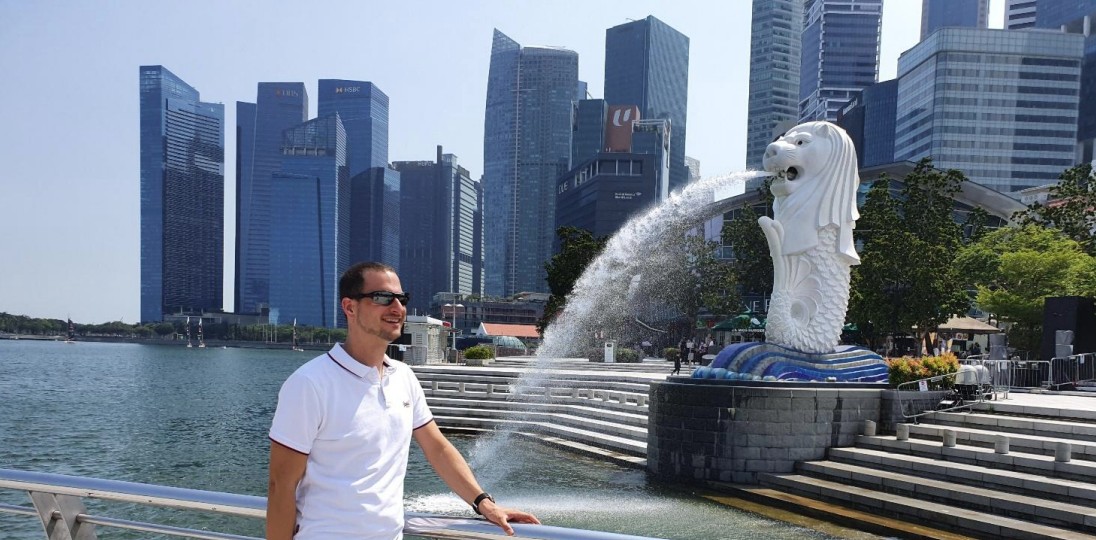 A man stands in front of a stone statue, with skyscrapers in the background.