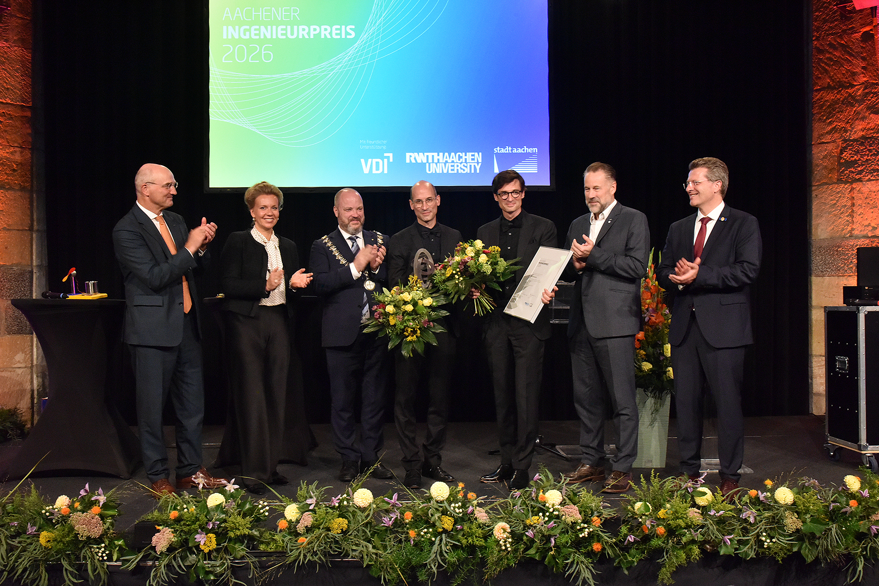 Auf dem Gruppenbild auf der Bühne sind die relevanten Personen rechts und links der beiden Preisträger beim gemeinsamen Applaus zu sehen.