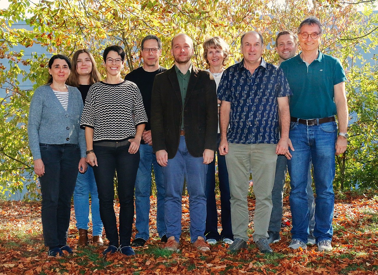 Gruppenfoto eines gemischten Teams aus Frauen und Männern im Außenbereich. Vor einem Hintergrund aus herbstlich gefärbten Pflanzen und Laub auf dem Boden steht der neue Lehrstuhlleiter in der Mitte, umgeben von seinen lächelnden wissenschaftlichen und administrativen Mitarbeiter:innen.