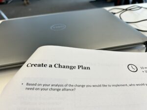 Papier auf dem steht: "Create a Change Plan- Based on your analysis of the change you would like to implement who would" Der Rest der Zeile ist außerhalb des Bildausschnitts. In der nächsten Zeile steht: "need on your change alliance?"