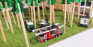 Ein aufgebauter Wald aus Holzbäumen mit Spielzeug-Feuerwehrautos dazwischen auf einer grünen Wiese