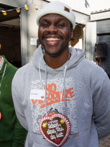 A man with a gingerbread heart hanging around his neck smiles at the camera.