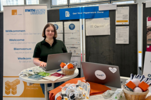 Eine Frau, die bei einer Veranstaltung hinter einem Stand mit Laptops steht, umgeben von Informationsmaterialien und Werbegeschenken.