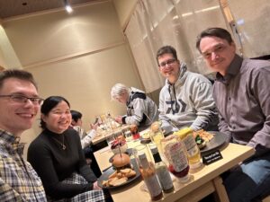 Four people are sitting at a table in a restaurant, smiling at the camera; there are burgers in front of them.