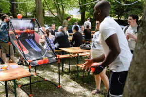 People at a summer party in the park, a man playing basketball at the shooting range, others sitting at tables.