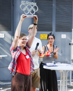 Andrea Bönsch hebt einen IT Center Pokal mit olympischen Ringen hoch, neben zwei Kollegen bei einem Außenevent mit Stehtischen.