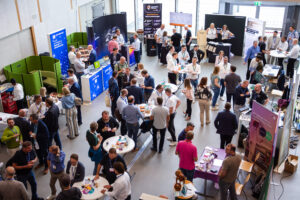 A bird's-eye view of a crowded exhibition hall with many booths, information boards, and groups of people engaged in conversation.