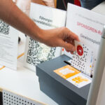 Hand wirft eine Gewinnspielkarte in eine schwarze Box, im Hintergrund Infotafeln mit QR-Codes und deutschen Texten.