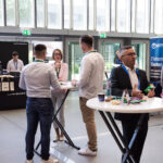 Menschen im Foyer einer Konferenz im Gespräch an Stehtischen, mit Firmenständen und Roll-up-Bannern im Hintergrund.