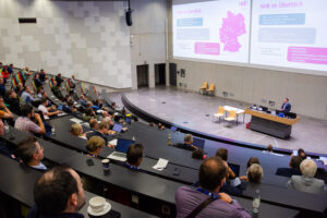 A large lecture hall filled with people sitting at their laptops, listening to a speaker standing in front of a screen showing a presentation that provides an overview of NHR.