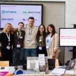Five smiling employees stand arm in arm in front of two monitors displaying partner logos.
