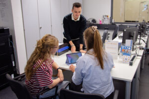 Two women are sitting at a table with a laptop; in front of them stands a man leaning forward