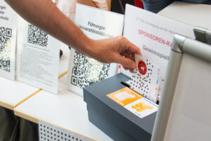 Das Bild zeigt eine Hand welche eine Karte hält und diese gerade in eine Gewinnspielbox geworfen wird.