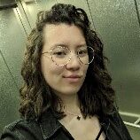 Porträt einer jungen Frau mit langen dunklen und lockigen Haaren. Sie trägt eine Brille und eine schwarze Jeansjacke